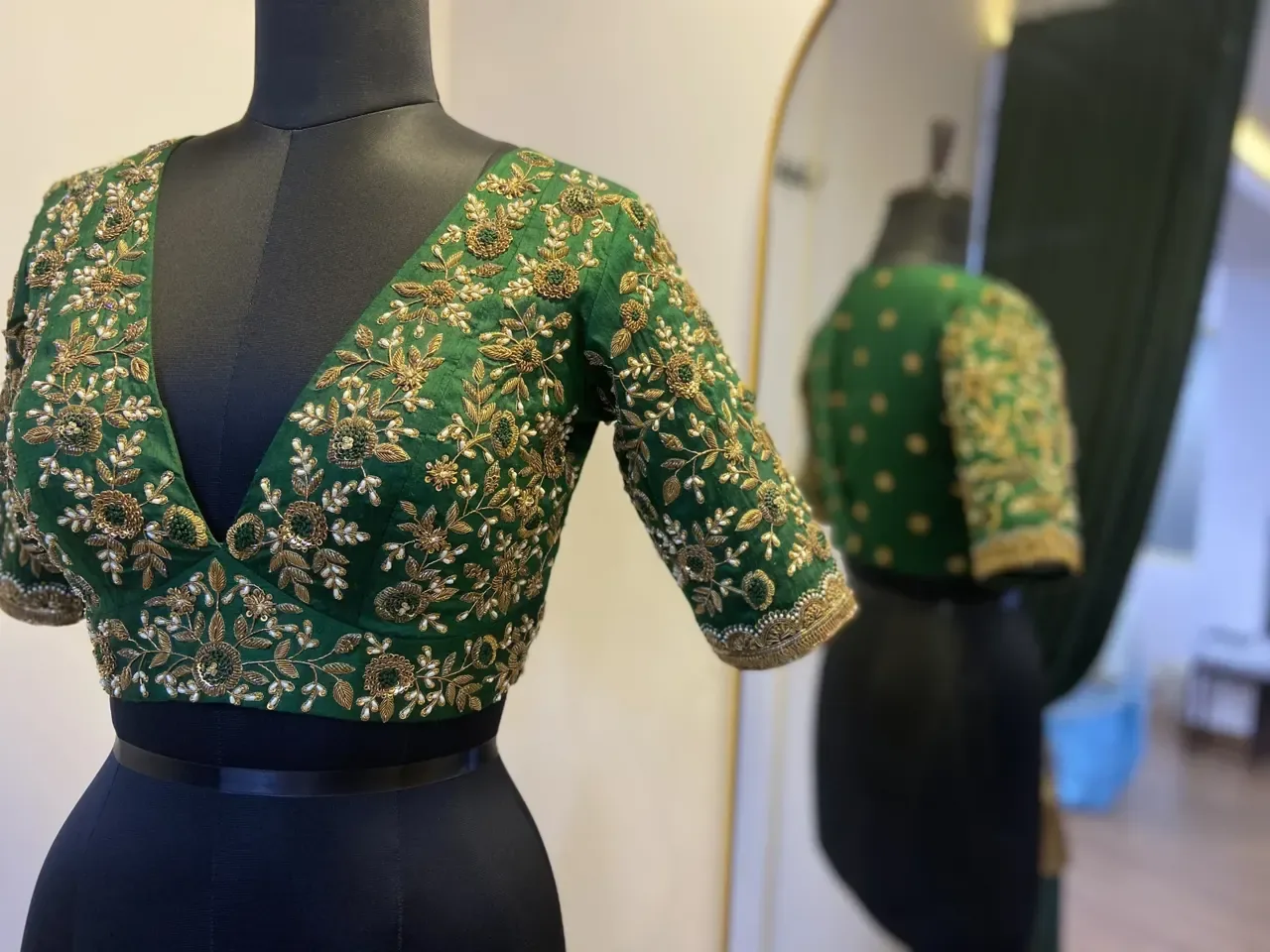 Green blouse with floral embroidery on a dress form, reflected in an arched mirror at Atelier Nur.